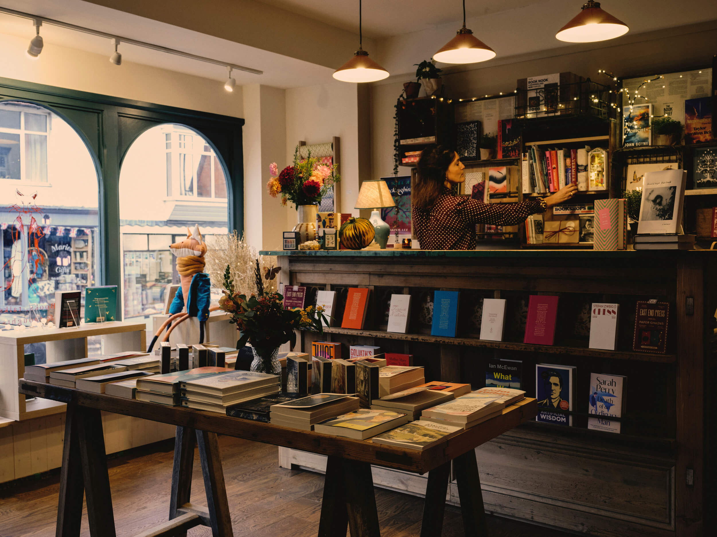 Interior shelves at Fox & King, Independent Bookshop, Tisbury, filled with carefully selected fiction and nonfiction arranged for browsing and discovery. Chloe King places a book on a shelf.