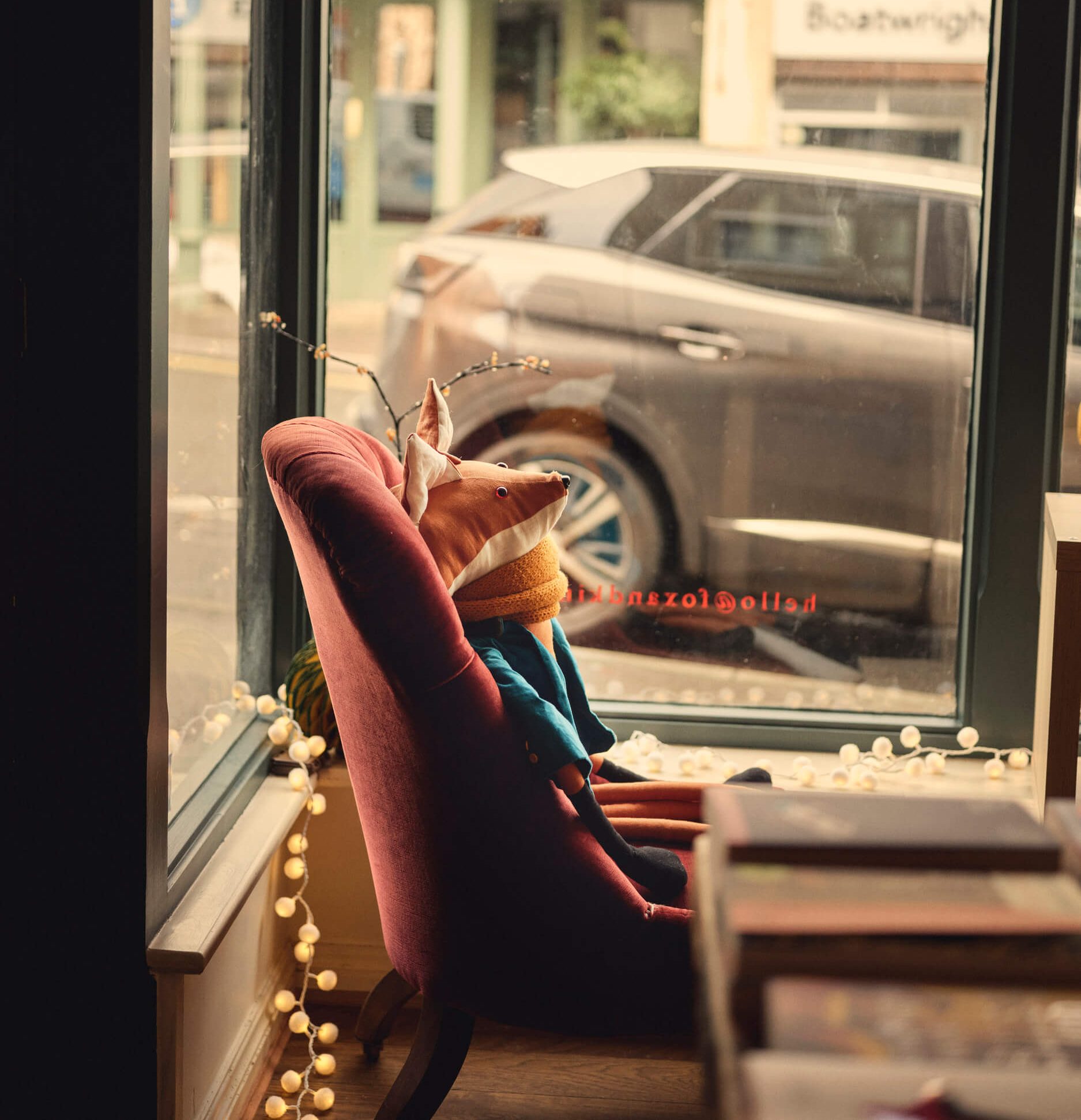 A soft toy Fox sits in a plush red velvet chair in the window display at Fox & King bookshop in Tisbury. He looks out into the street from the window. he is wearing a blue jacket and a yellow scarf.