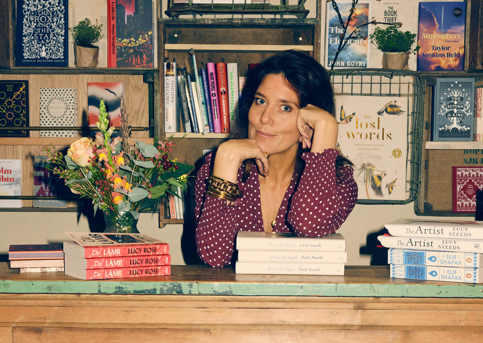 A woman, Chloe King, in a maroon polka dot shirt sits at a desk with her chin resting on her hands, surrounded by books and a bouquet of flowers, with a bookshelf in the background at Fox & King bookshop in Tisbury